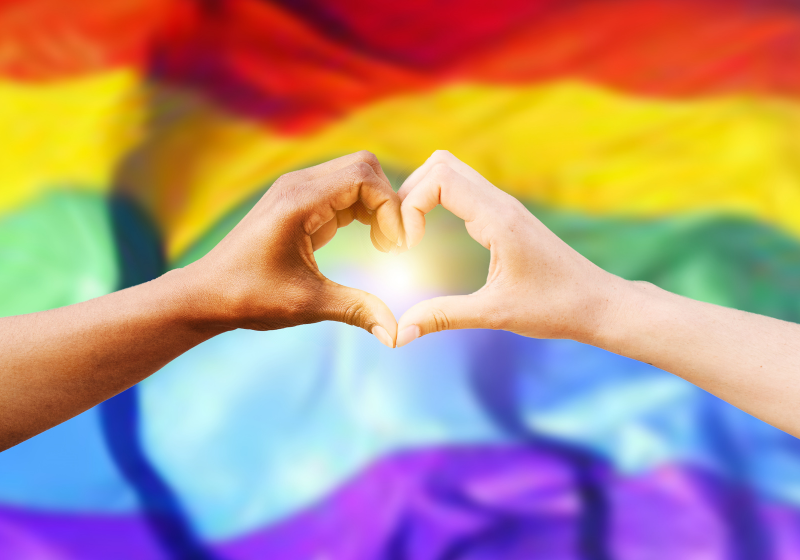 A heart shape made by hands in front of a rainbow flag, conveying a message of love and diversity.