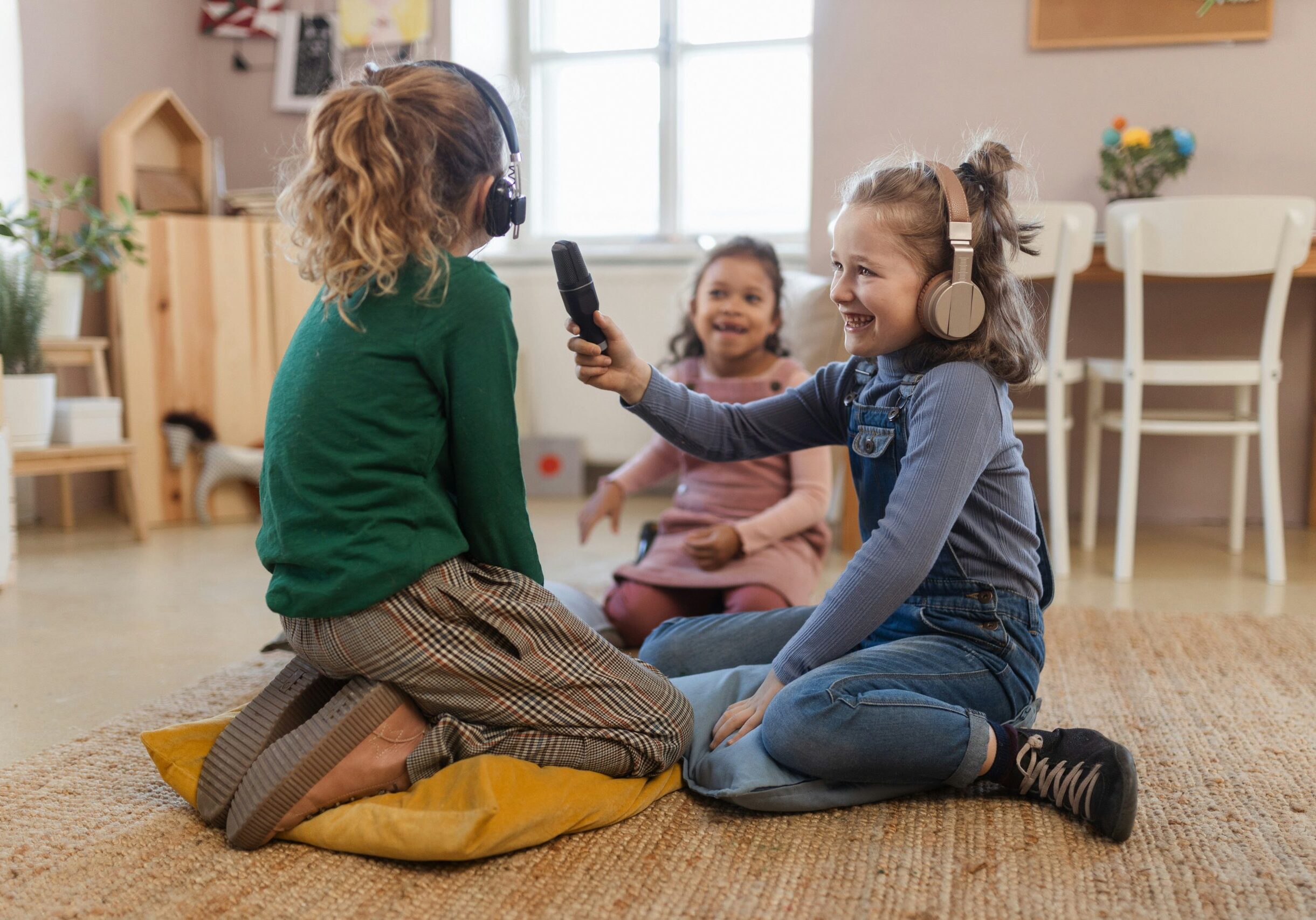 3 happy girls, 2 with headphones and a microphone playing at interviewing, with a third watching.