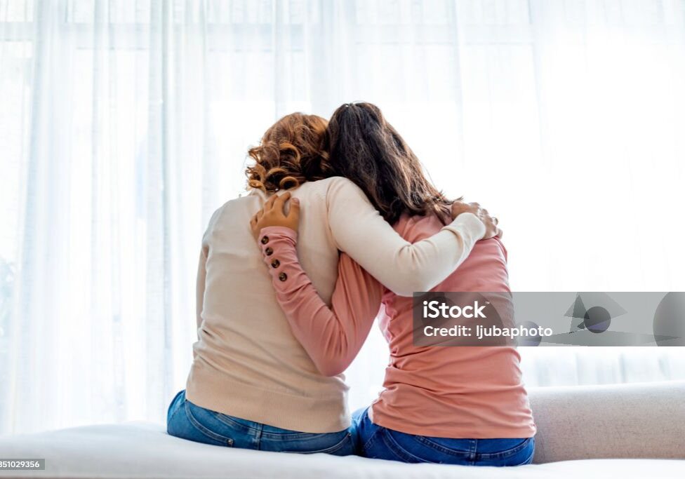 Rear back view of a mother and daughter embrace sitting on bed at home, older sister consoling younger teen, girl suffers from unrequited love share secrets trustworthy person relative people concept